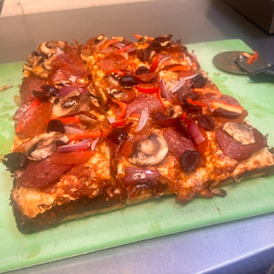 A square Italian pizza with pepperoni, mushrooms, red onions, and olives on a green cutting board.