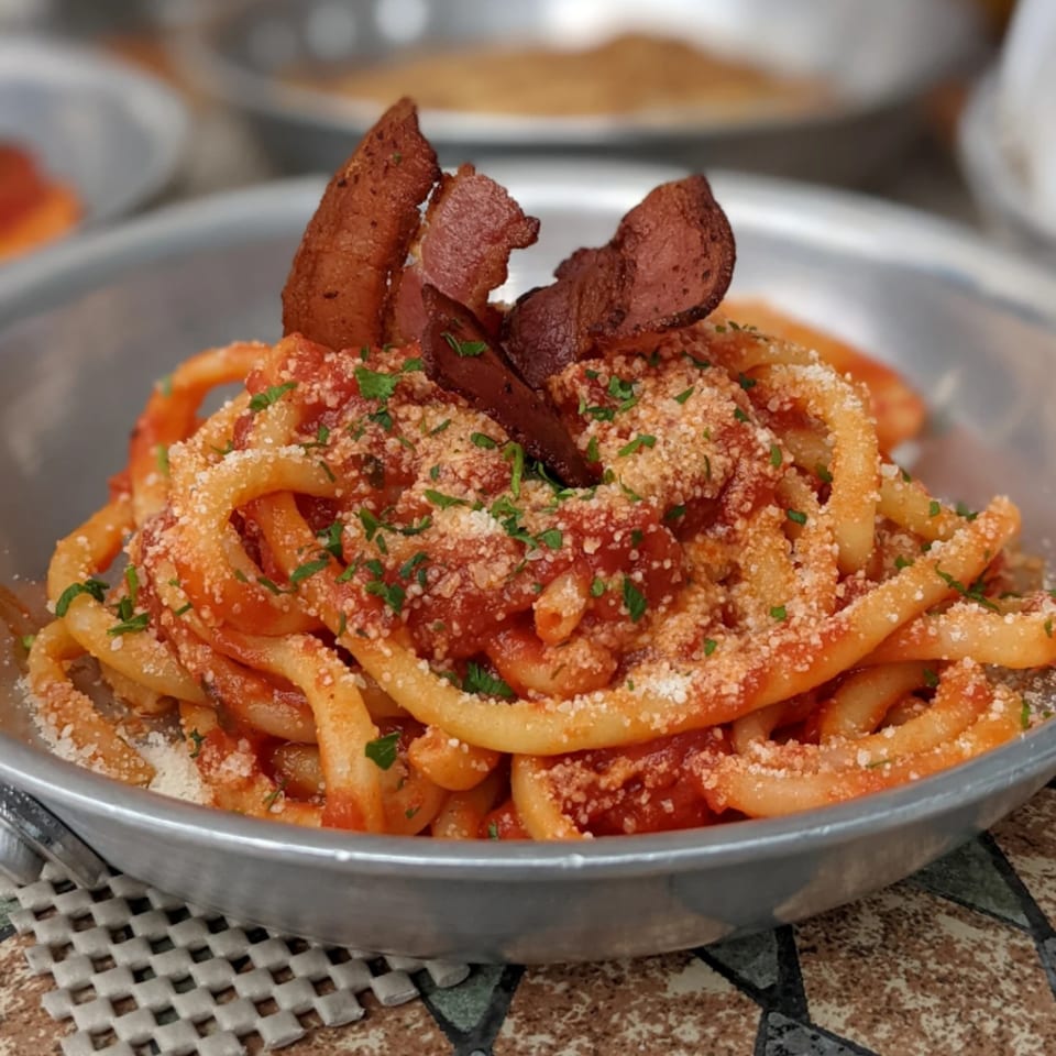 Bucatini pasta with red sauce, parmesan cheese, parsley, and crispy bacon in a metal bowl.